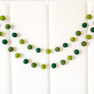May include: A garland of green felt balls strung on white thread. The balls are arranged in a gradient from dark green to light green.