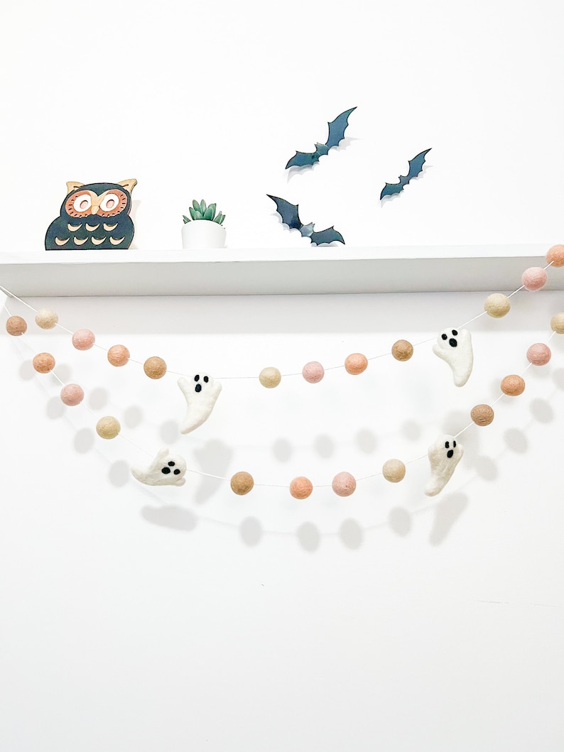 May include: A Halloween garland with white felt ghosts and peach and brown felt balls. The garland is hanging on a white shelf with a black owl figurine and a small potted plant.