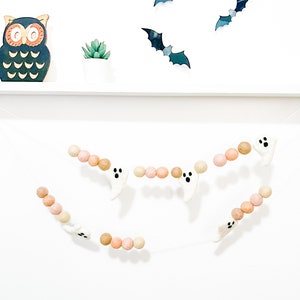 May include: A Halloween garland with white felt ghosts and brown and pink felt balls. The garland is hanging on a white wall with a black owl and bats on a shelf above.
