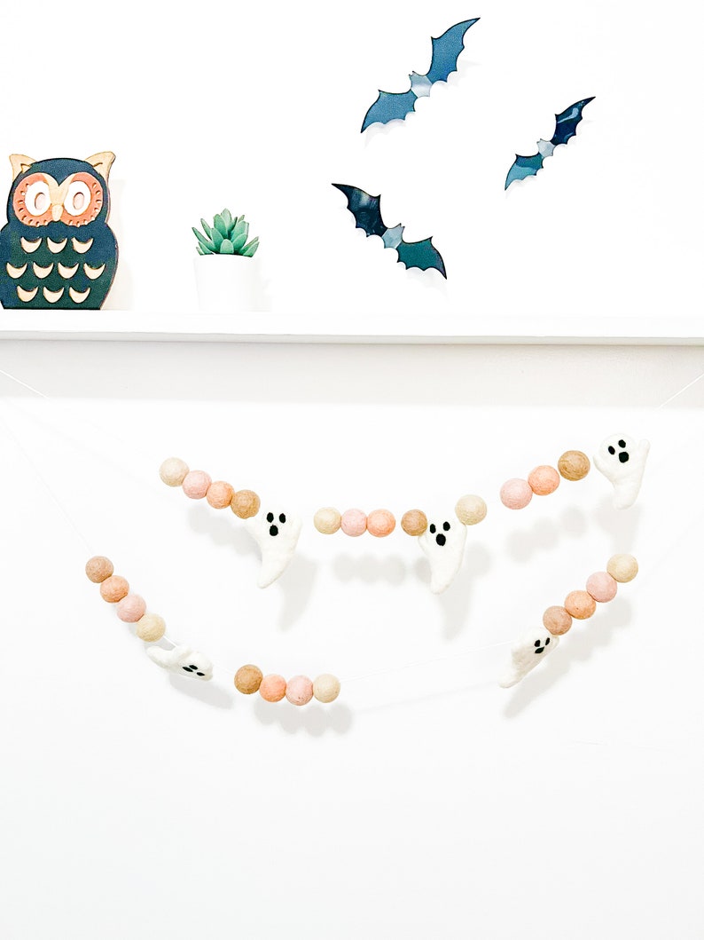 May include: A Halloween garland with white felt ghosts and brown and pink felt balls. The garland is hanging on a white wall with a black owl and bats on a shelf above.