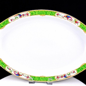 Könnte beinhalten: Weiße ovale Servierplatte mit grünem und goldenem Rand. Der Rand weist ein Blumen- und Vogeldesign mit roten, blauen und gelben Akzenten auf. Die Platte hat eine glatte, glänzende Oberfläche und wird von transparenten Kunststoffständern getragen.