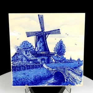 May include: A blue and white ceramic tile featuring a windmill scene. The artwork depicts a Dutch landscape with a windmill, houses, a bridge, and a waterway. The tile is square and has a glossy finish.