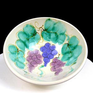 May include: A white ceramic bowl with a hand-painted grape design. The bowl features clusters of purple and blue grapes, surrounded by green leaves and vines. The rim is a simple, rounded edge.