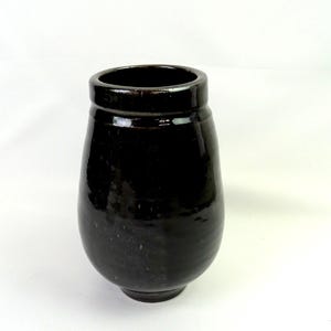 Puede incluir: Un jarrón de cerámica de color marrón oscuro brillante con un cuerpo redondeado y bulboso y un borde grueso ligeramente ensanchado. La superficie del recipiente presenta un acabado liso y reflectante que capta la luz, resaltando el color profundo y uniforme del esmalte.