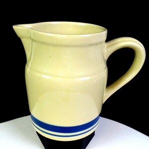 May include: A cream-colored ceramic pitcher featuring a smooth, glossy finish and a rounded handle. The base of the pitcher is decorated with two distinct, horizontal dark blue stripes that wrap around the lower portion of the vessel.