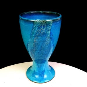 May include: A ceramic goblet featuring a vibrant blue glaze with a mottled, textured finish that transitions from a deep cobalt to a lighter turquoise hue. The vessel has a wide, open rim, a tapered body, and a circular pedestal base.