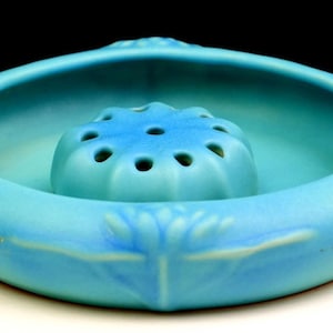 May include: A round, matte turquoise ceramic flower frog bowl featuring a central dome with multiple circular openings for stems. The exterior rim is decorated with embossed floral motifs, and the overall finish is a solid, vibrant blue-green.