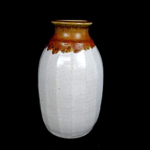 Puede incluir: Un jarrón de cerámica con cuerpo blanco y borde marrón. El jarrón tiene una superficie texturizada y una abertura ligeramente ensanchada. El borde marrón tiene un borde más oscuro y texturizado. El jarrón está sobre un fondo negro.