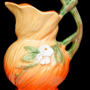 May include: A ceramic pitcher featuring a gradient color scheme transitioning from a light yellow at the top to a vibrant orange at the base. The surface has a textured, swirling pattern and is decorated with a raised white flower motif.