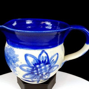 Puede incluir: Una jarra de cerámica con un diseño floral azul y blanco. La parte superior y el asa son de un azul liso y brillante, mientras que el cuerpo presenta una flor azul pintada a mano y follaje sobre un fondo blanco. La jarra tiene un pico.