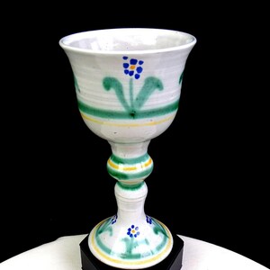 May include: A white ceramic goblet featuring hand-painted green horizontal bands, yellow accent lines, and small blue floral motifs with yellow centers. The vessel has a flared rim, a rounded stem, and a circular base, all decorated with matching patterns.