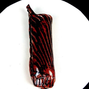 May include: A handmade ceramic vase with a glossy finish. The vase is dark red with black diagonal stripes, creating a textured look. The top has an irregular shape. The vase is approximately 15 cm tall.