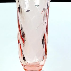 May include: A pink glass vase with a flared top and a round base. The vase has a twisted design, with vertical lines and a subtle color gradient. The glass appears to be smooth and transparent, allowing light to pass through. The vase is likely used for decorative purposes.