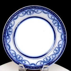 Puede incluir: Un plato de cerámica blanca con un borde decorativo azul y dorado. El plato presenta un detallado diseño floral y de volutas azules alrededor del borde. El plato se exhibe sobre un soporte transparente sobre un fondo negro.