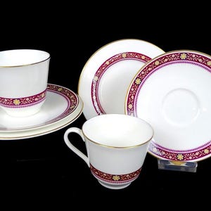 Peut inclure: Service à thé blanc avec bordure dorée et motif floral bordeaux et or. L'ensemble comprend deux tasses et soucoupes, et deux soucoupes supplémentaires. Les tasses sont sur fond noir.