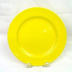 May include: A solid, bright yellow circular plate with a smooth, glossy finish and a wide, flat rim. The plate is positioned upright on two small, clear plastic display stands, showcasing its vibrant, uniform color and simple, clean design.