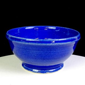 Puede incluir: Un cuenco de cerámica de color azul cobalto profundo y vibrante con un acabado liso y brillante. El cuenco presenta un cuerpo redondeado que se estrecha hacia una base circular, con un borde distintivo y sutiles estrías horizontales.