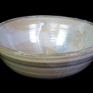 May include: A round ceramic bowl with a glossy, off-white interior and a speckled, light tan exterior. The bowl features subtle horizontal ridges along the outer wall and a smooth, wide rim, showcasing a handcrafted appearance with visible glaze variations.