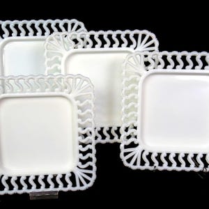 May include: A set of four square white milk glass plates featuring a decorative openwork lattice border with a repeating wave pattern. Each plate has a smooth, solid center and a raised, intricate edge design that frames the square surface.