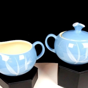 May include: A light blue ceramic creamer and sugar bowl set. The creamer has a spout and handle, while the sugar bowl has a lid and two handles. Both pieces feature a white leaf design. The set is displayed on a black surface.