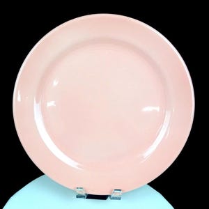 May include: A round, solid light pink ceramic dinner plate with a smooth, glossy finish and a wide, flat rim. The plate features a simple, clean design with a uniform pastel colour throughout the entire surface, presenting a classic circular shape.