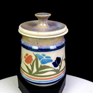 May include: A ceramic jar with a matching lid, featuring a cream-colored base with a hand-painted floral pattern in orange, blue, and dark blue, accented by green leaves and horizontal stripes at the bottom, finished with a blue and tan glaze.