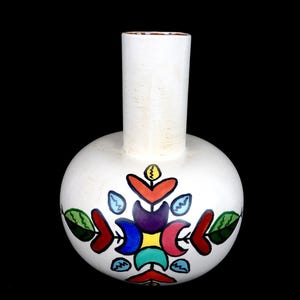 May include: A white ceramic vase with a rounded body and a cylindrical neck. The front features a colourful, symmetrical hand-painted design consisting of red, orange, yellow, purple, blue, teal, and green leaf and petal-like shapes.