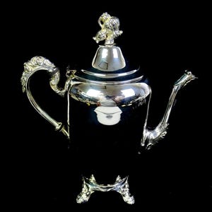 May include: A polished silver teapot featuring a smooth cylindrical body, a curved handle with ornate detailing, a matching spout, and a lid topped with a decorative finial. The teapot stands on three small, intricate, and ornate feet.