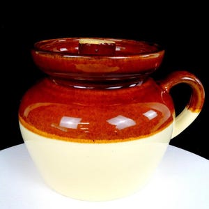 Op de afbeelding: Een keramische pot met een glanzend bruin geglazuurd bovengedeelte en een crèmekleurig onderlichaam. Het vat heeft een wijde opening, een bijpassend bruin deksel met een centrale knop en één gebogen handvat aan de zijkant voor grip.