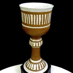 May include: A ceramic goblet featuring a tall, slender stem and a wide bowl. The piece is finished in a warm, earthy brown glaze with contrasting white vertical stripes painted around the rim, the center of the stem, and the circular base.