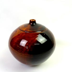 May include: A spherical ceramic vase with a glossy, dark brown glaze that transitions into lighter amber tones. The vessel features a smooth, rounded body and a small, narrow circular opening at the top, reflecting light on its polished surface.