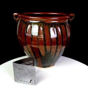 Op de afbeelding: Een handgemaakte keramische pot met een glanzende afwerking. De pot heeft verticale strepen in bruin- en zwarttinten, met twee kleine handvatten. Een gevouwen kaart met tekst en een cirkelvormig ontwerp staat voor de pot.