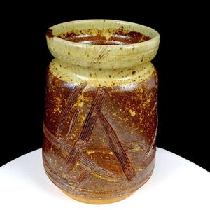 Può includere: Un vaso cilindrico in ceramica con un bordo largo e spesso. L'esterno presenta una smaltatura maculata marrone e marrone chiaro con un motivo strutturato di linee incise che si incrociano sul corpo. La base è di colore marrone chiaro non smaltato.