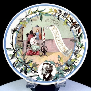 Puede incluir: Un plato decorativo que presenta una escena de "Der fliegende Holländer". El plato tiene un borde azul y blanco con una ilustración central de una mujer, una rueca y notas musicales. También se incluye un retrato de un hombre.