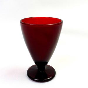 May include: A deep red glass goblet featuring a smooth, rounded bowl that tapers towards a short, thick stem and a circular base. The vessel is crafted from translucent, solid-coloured red glass with a polished finish and a simple, clean silhouette.