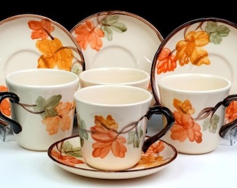 May include: A set of three cream-coloured ceramic cups and saucers featuring a painted pattern of orange and green autumn leaves. Each cup has a dark brown handle and a matching saucer, with the leaf design repeated on both the cups and saucers.