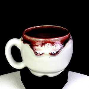 Puede incluir: Una taza de cerámica blanca con cuerpo redondeado y asa. El borde es de color burdeos oscuro, con un efecto de goteo blanco y rojo. El interior de la taza es de color burdeos oscuro. La taza está sobre un fondo negro.