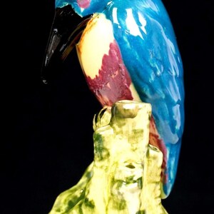 May include: A ceramic kingfisher figurine perched on a textured, moss-green tree stump. The bird features a glossy bright blue head and back, a cream-colored chest, and a dark maroon patch on its side, finished with a long, pointed black beak.