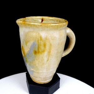May include: A handmade ceramic mug with a rounded handle. The mug has a beige base color with streaks of brown and blue. The mug is displayed on a black stand against a black background.