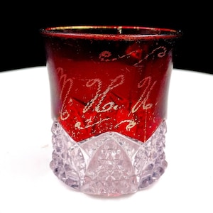 Pode incluir: Um copo com uma secção superior vermelho-profundo com letras cursivas gravadas e uma base inferior transparente e texturizada com um padrão de diamante. O rebordo tem uma banda vermelha metálica, e o vidro mostra um contraste entre o topo liso e colorido e a base facetada e transparente.