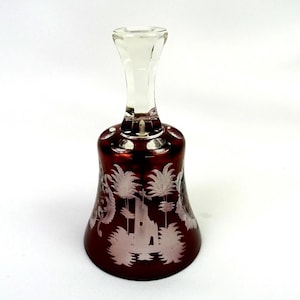 Peut inclure: Une cloche en verre rouge foncé avec une poignée en verre transparent et des motifs blancs gravés représentant des palmiers et une structure de bâtiment. La cloche présente une base évasée et une finition lisse et polie, contrastant les couleurs.