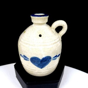 May include: A small, off-white ceramic jug with a speckled texture, featuring a hand-painted blue heart design on the front. The jug has a single side handle, a narrow neck with a blue rim, and a small circular hole located just below the neck.