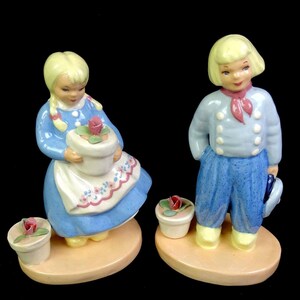 Op de afbeelding: Twee porseleinen beeldjes van kinderen met blond haar op ovale voetstukken. Eén figuur draagt een blauwe jurk met een wit schort en houdt een kleine bloempot met een roos vast. De andere figuur draagt een lichtblauw jasje, een rode sjaal en een blauwe broek.