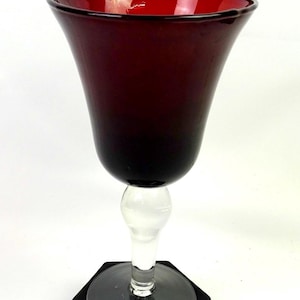 May include: A deep red glass goblet featuring a wide, flared bowl that transitions into a clear, spherical stem and a circular base. The glass displays a rich, dark crimson hue throughout the upper portion, contrasting with the transparent stem.