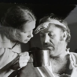 以下が含まれることがあります： 女性が男性に金属製のカップを差し出している白黒写真。男性は頭にタオルを巻き、口ひげを生やしています。女性はカップを見下ろしています。