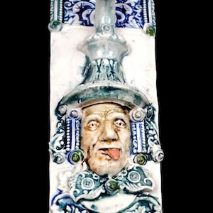 May include: A vertical ceramic wall plaque featuring a relief sculpture of a winged figure at the top and a central face with a protruding tongue. The piece is glazed in shades of white, blue, and green with intricate scrollwork and floral motifs.
