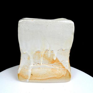 May include: A handmade, rectangular ceramic vase with a textured, off-white glaze. The glaze has drips and streaks of tan and brown, adding a rustic, organic feel. The vase sits on a white surface against a black background.