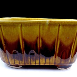 May include: A rectangular ceramic planter with a ribbed design, featuring a gradient of colors from golden yellow to deep brown. The planter rests on three small feet and is suitable for indoor or outdoor use.