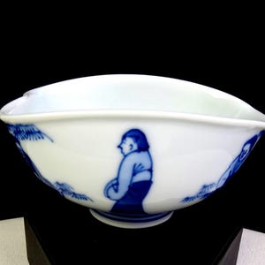 Puede incluir: Cuenco de cerámica blanca con un diseño pintado en azul. El cuenco presenta un borde curvo y una base con pie. El diseño azul representa figuras y follaje. El cuenco es probablemente para servir o exhibir.