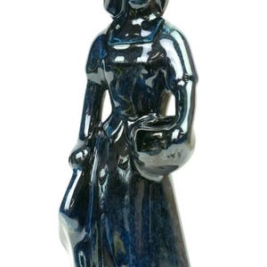 Puede incluir: Una figura de cerámica brillante de una persona con un tocado y un vestido largo, sosteniendo un cuenco pequeño en una mano. La escultura presenta un esmalte moteado en azul profundo y negro con reflejos brillantes en toda la superficie.